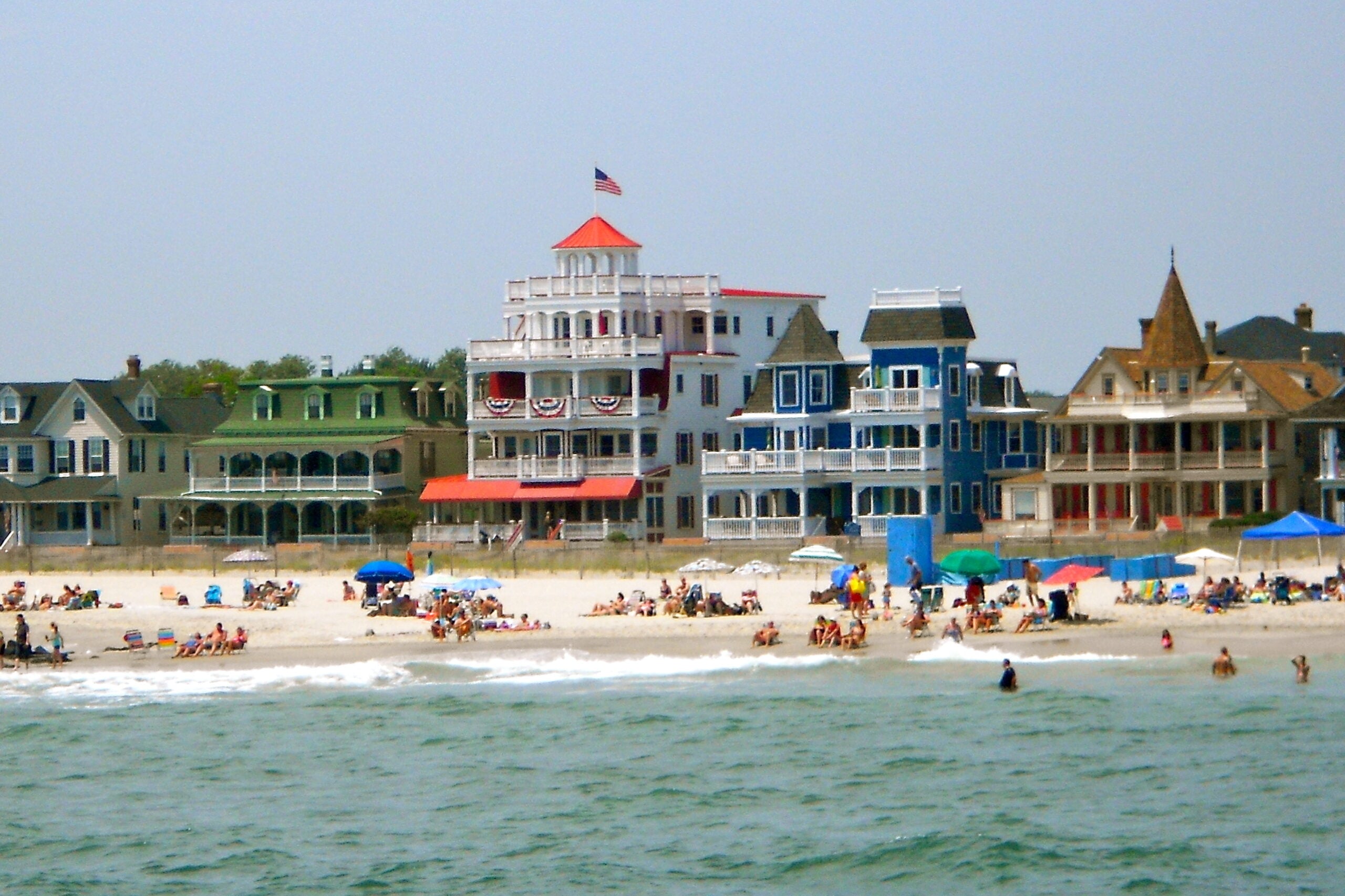 Colorful Victorian-style houses line a busy beach, with people relaxing under umbrellas and swimming in the ocean. An American flag flies atop the central building, and the sky is clear.