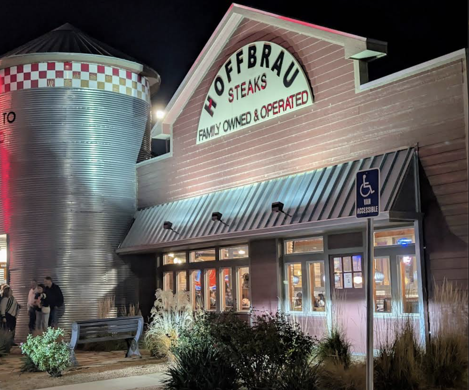 Exterior of Hoffbrau Steaks restaurant at night, with a large sign reading "Family Owned & Operated." Lights illuminate the building. A metal silo and a wheelchair accessible parking sign are visible.