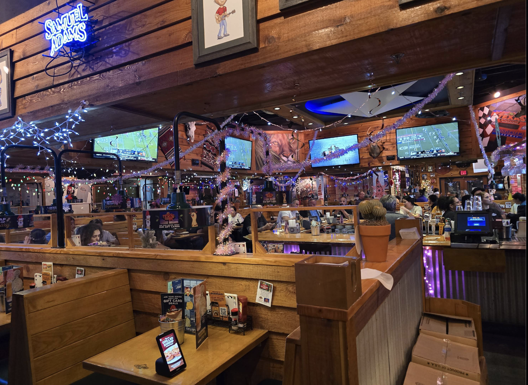 A lively restaurant with wooden booths and festive string lights, multiple TVs showing sports, people dining and talking, and a cactus plant on a divider. The atmosphere is warm and welcoming.