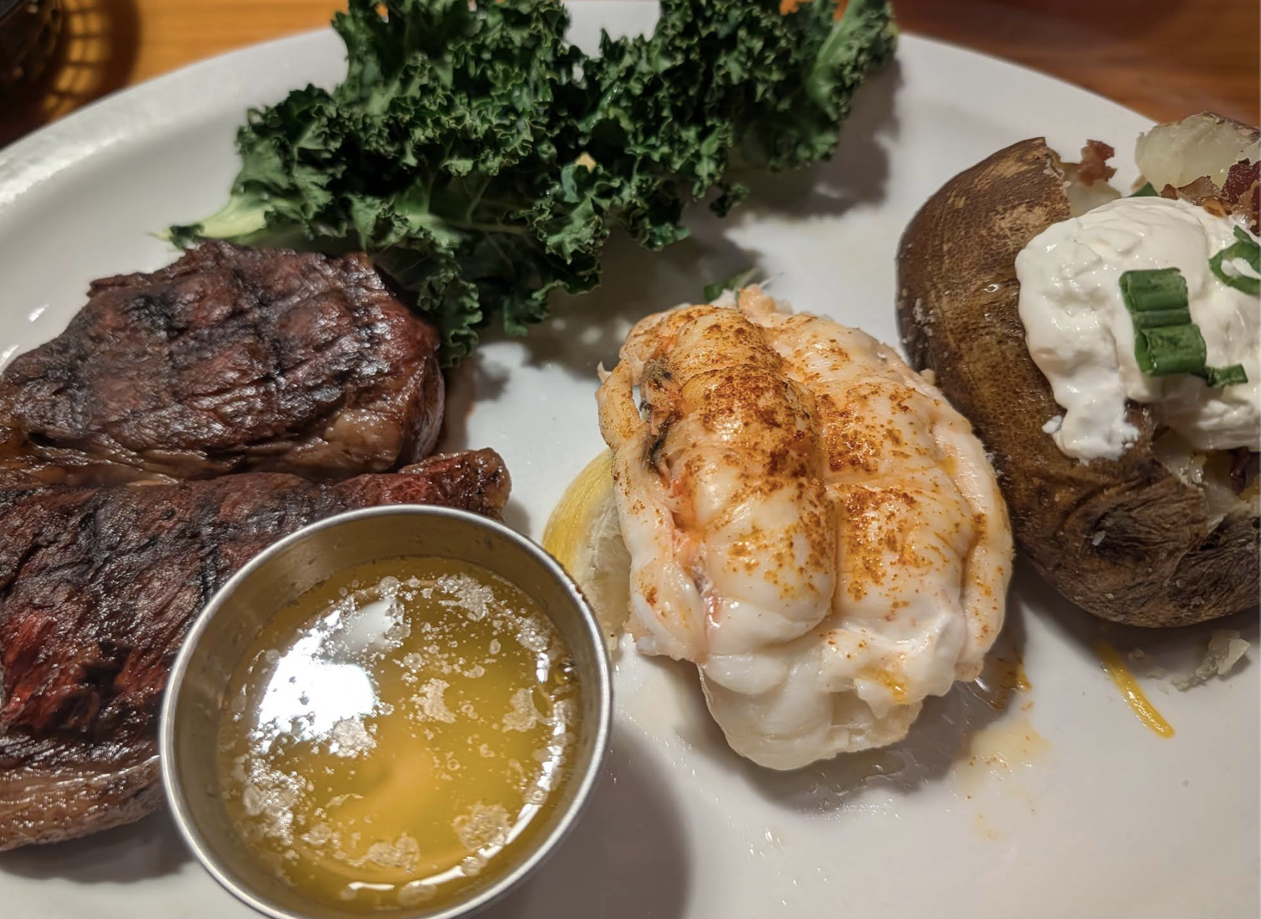 A dinner plate with grilled steak, a lobster tail sprinkled with paprika, a baked potato topped with sour cream and green onions, leafy kale garnish, and a small cup of melted butter.