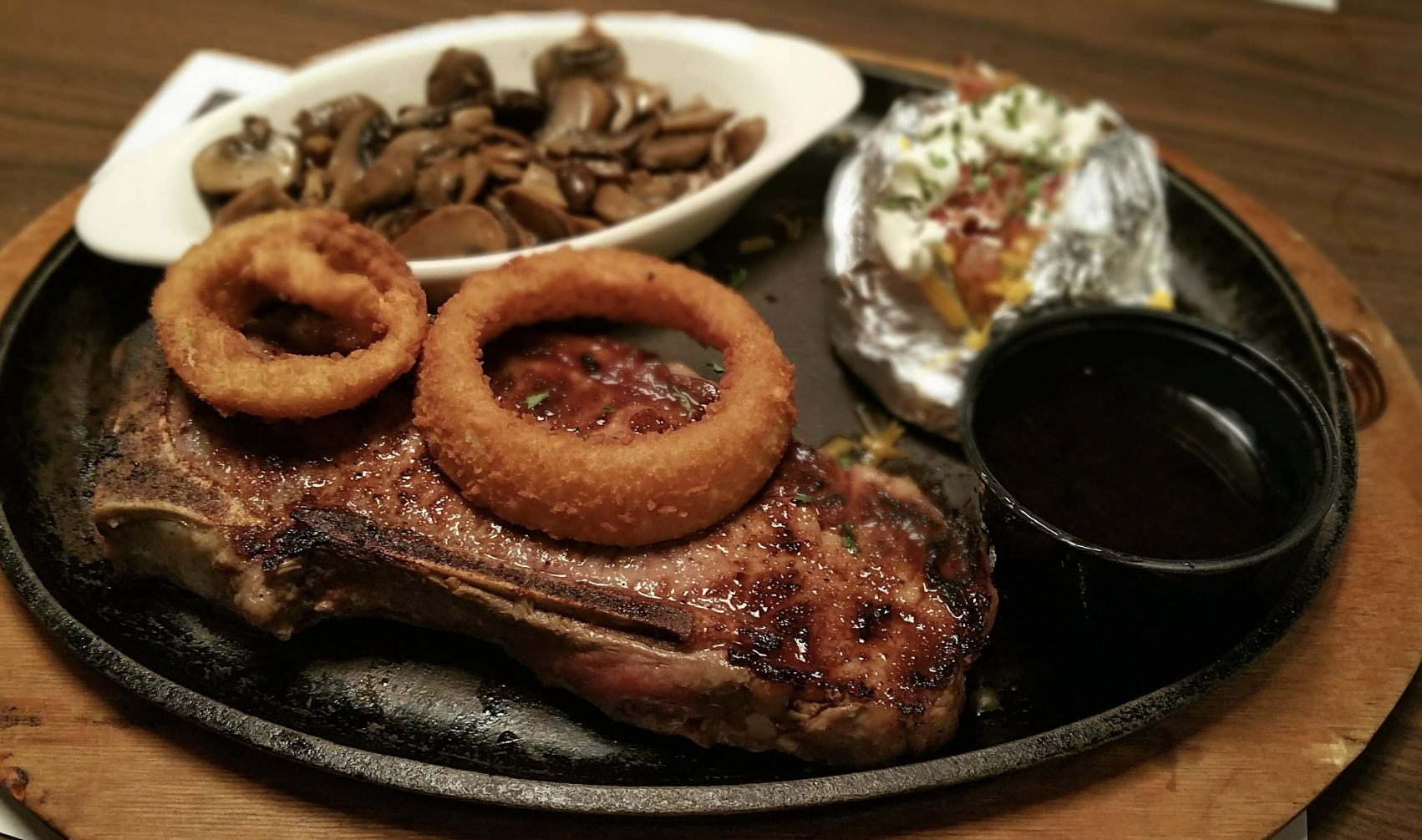 A grilled steak topped with two onion rings served on a hot plate, with a side of sautéed mushrooms, baked potato with toppings, and a small cup of sauce.
