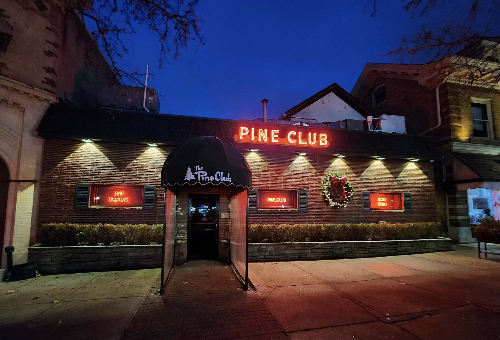 A brick building at dusk with a black awning reading "The Pine Club" and red neon signs that say "PINE CLUB." The windows display neon signs, and a holiday wreath hangs on the wall.