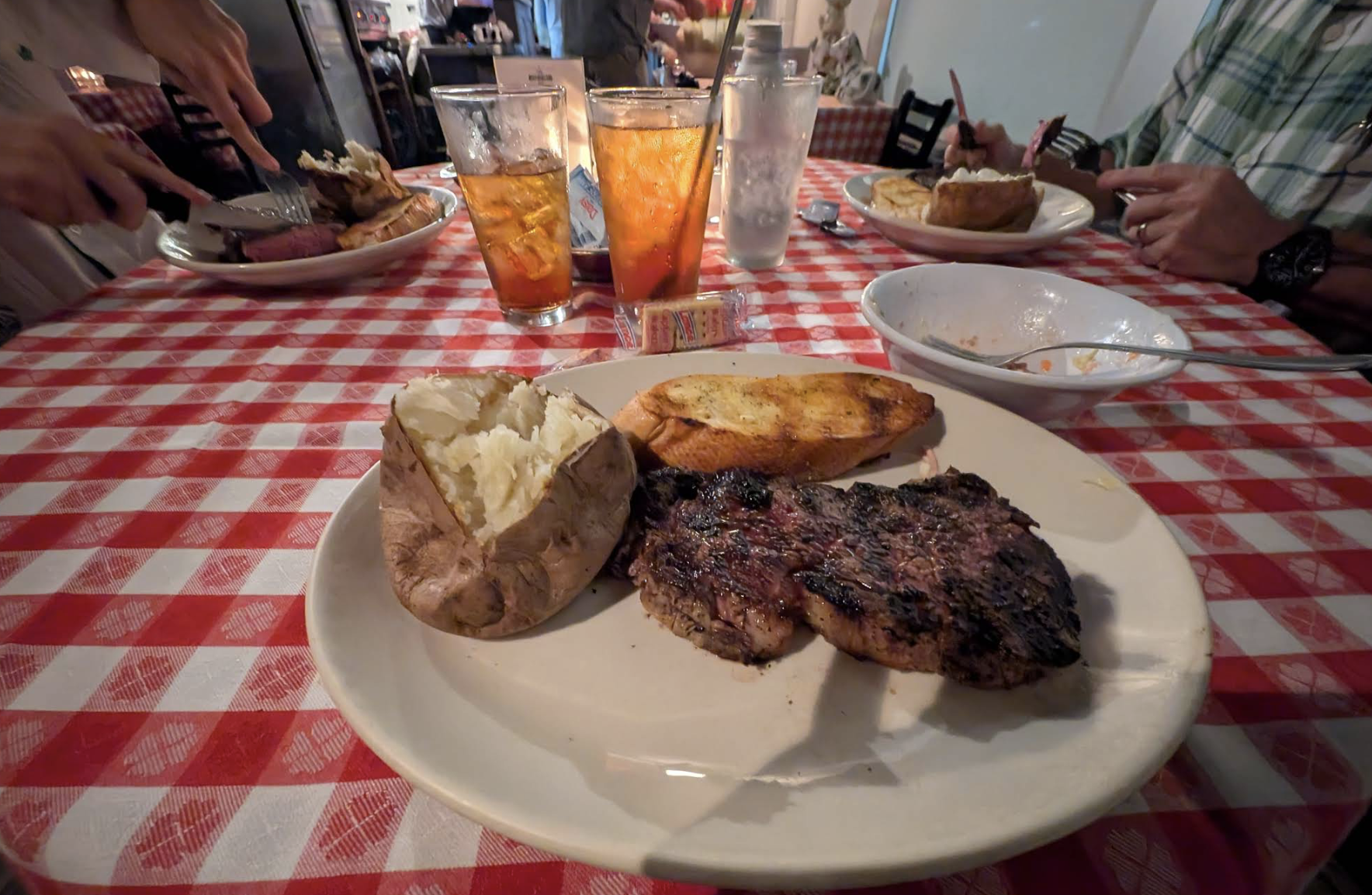 A dinner plate on a red checkered tablecloth holds a grilled steak, baked potato, and garlic bread. Three drinks, utensils, and plates with food are also on the table, with people dining in the background.