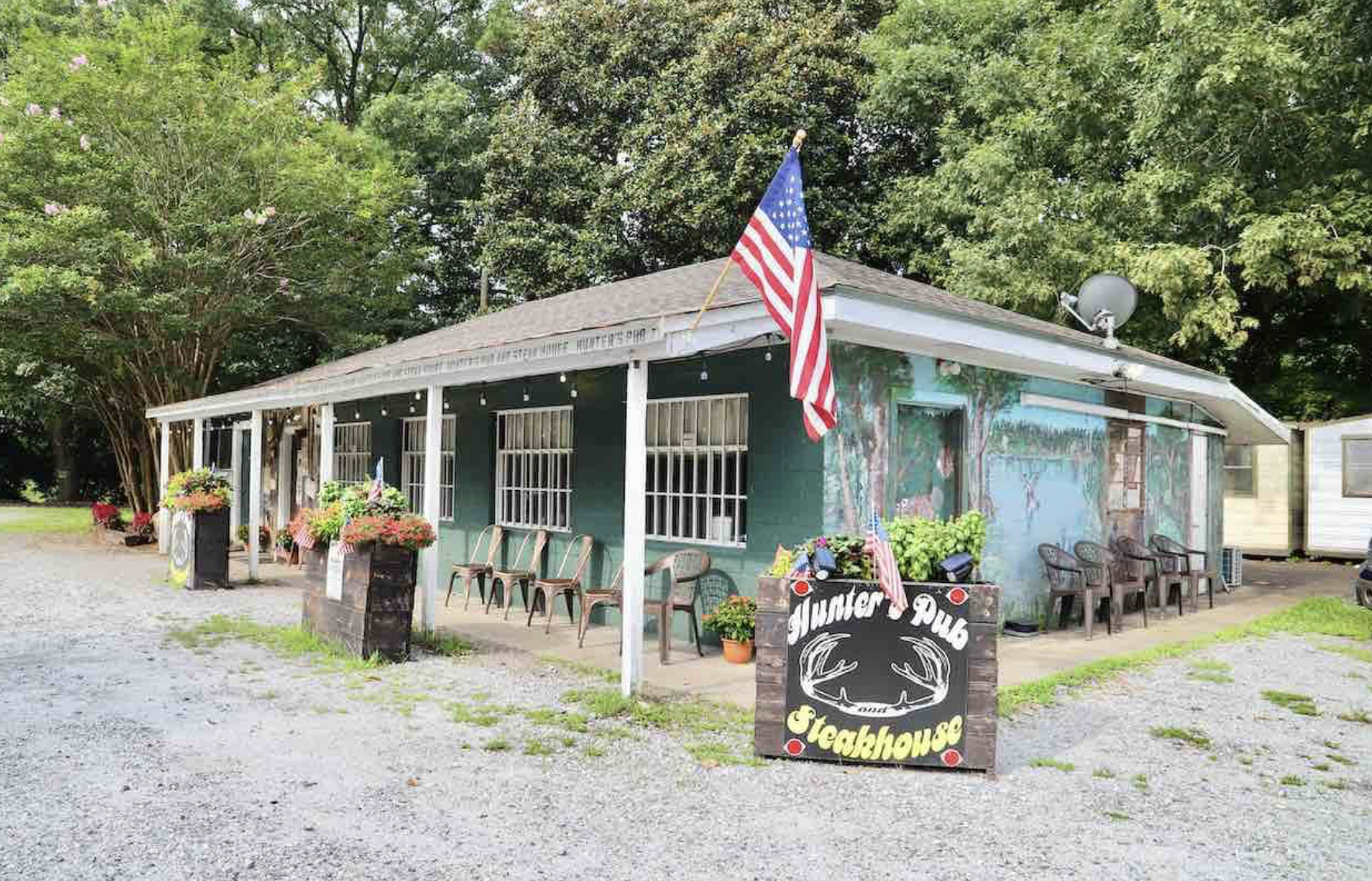 A rustic, single-story restaurant with an American flag, potted plants, and outdoor seating. A sign reads “Hunter Pub Steakhouse.” Surrounded by trees on a gravel lot under a bright sky.