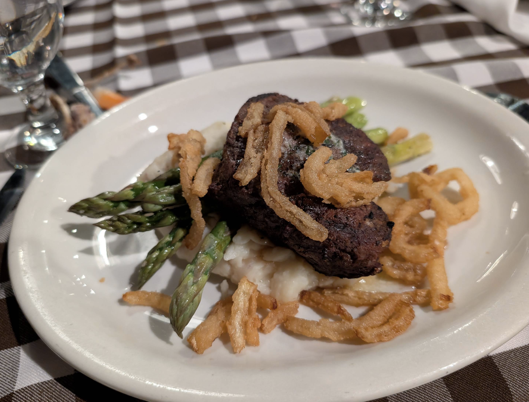 A plate with grilled steak on mashed potatoes, topped with crispy fried onions and served with asparagus spears, set on a brown and white checkered tablecloth.