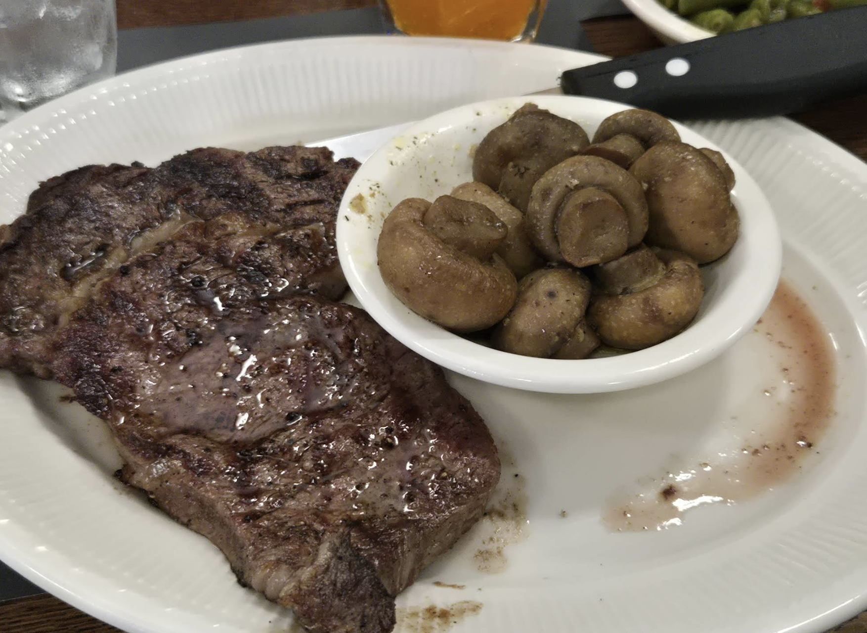 A plate with a cooked steak next to a small bowl filled with sautéed mushrooms. A steak knife rests on the plate, and some steak juices are visible.