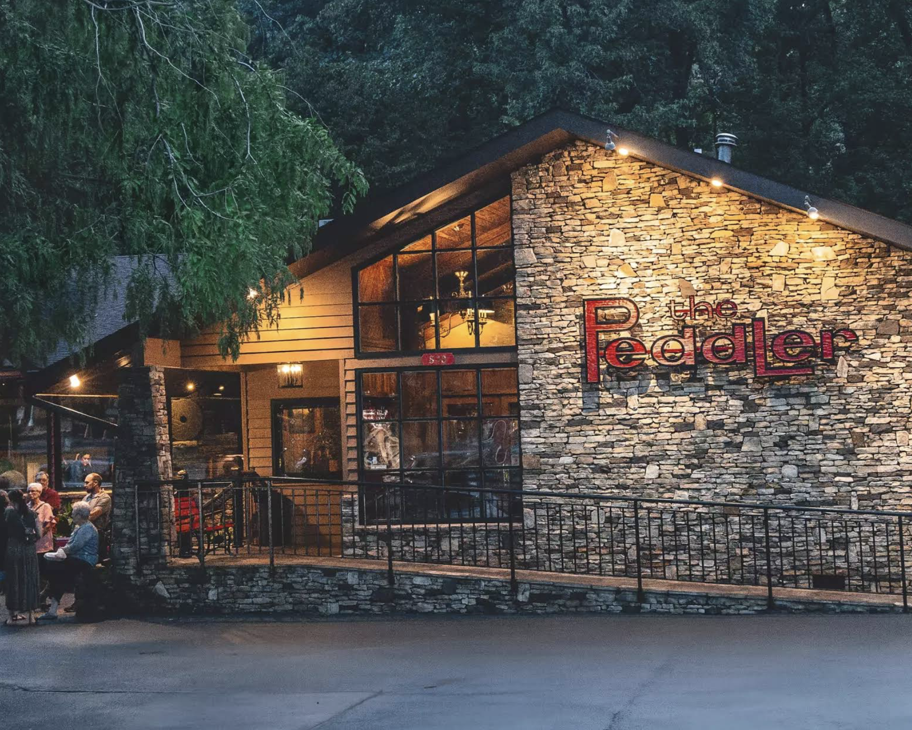 A rustic stone and wood building with large windows and a sign reading “The Peddler.” Several people are gathered outside under warm lights, surrounded by trees. The setting appears calm and inviting in the evening.