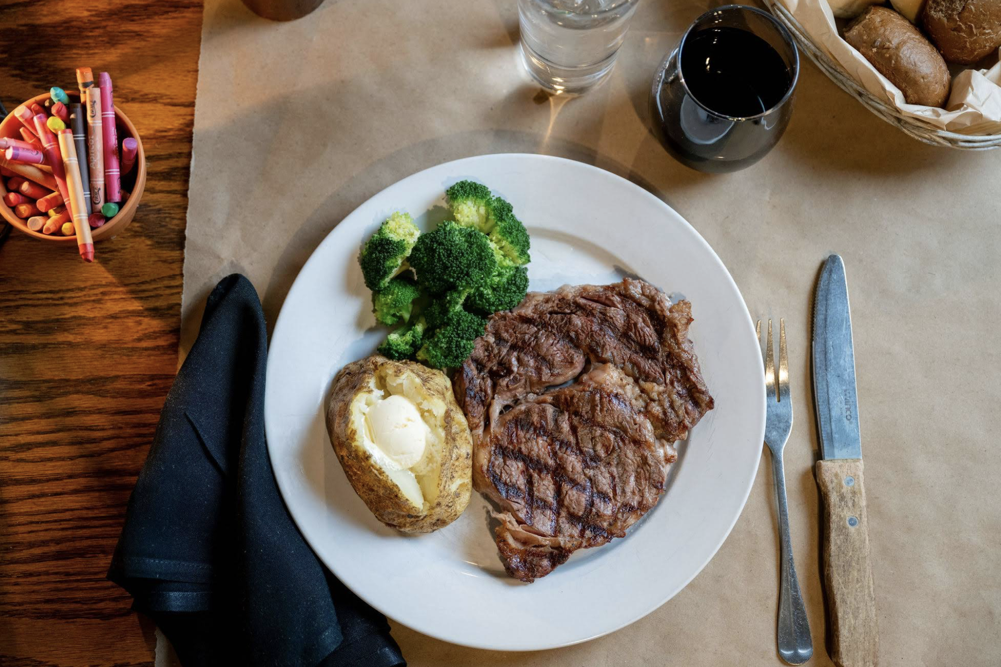 A grilled steak, baked potato with butter, and steamed broccoli are served on a white plate with a knife, fork, black napkin, glass of water, glass of red wine, bread basket, and a cup of crayons on a brown paper table cover.