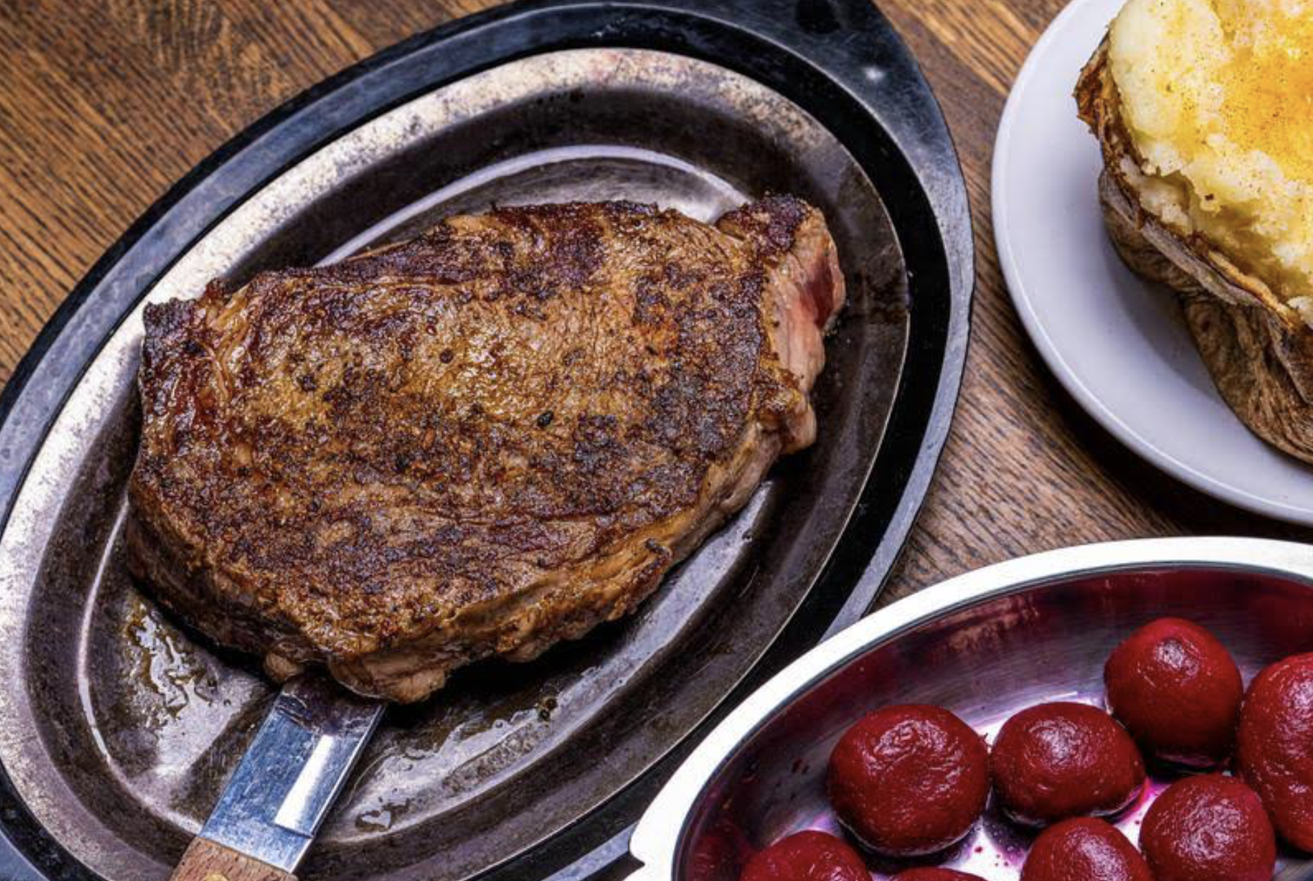 A grilled steak on a metal plate with a knife handle visible, next to a bowl of red beets and a baked potato topped with butter on a separate plate, all on a wooden table.