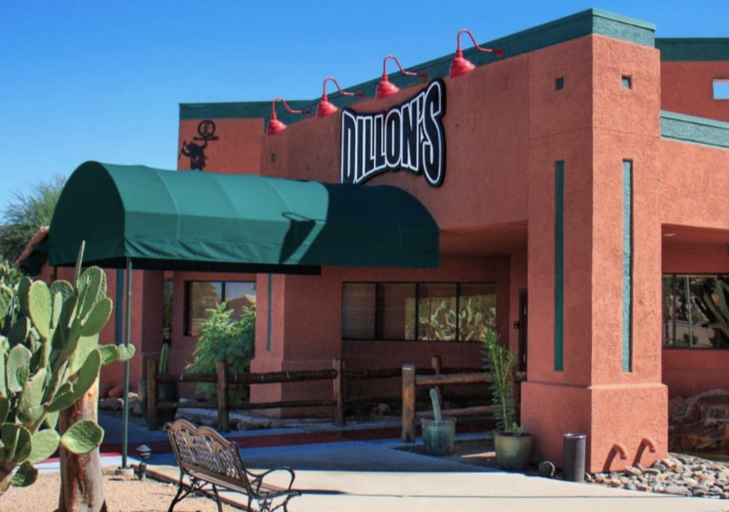 A southwestern-style restaurant with adobe-like orange walls, a green awning, and red light fixtures, featuring a sign that reads "Dillon’s." Desert plants and a bench are visible in front.