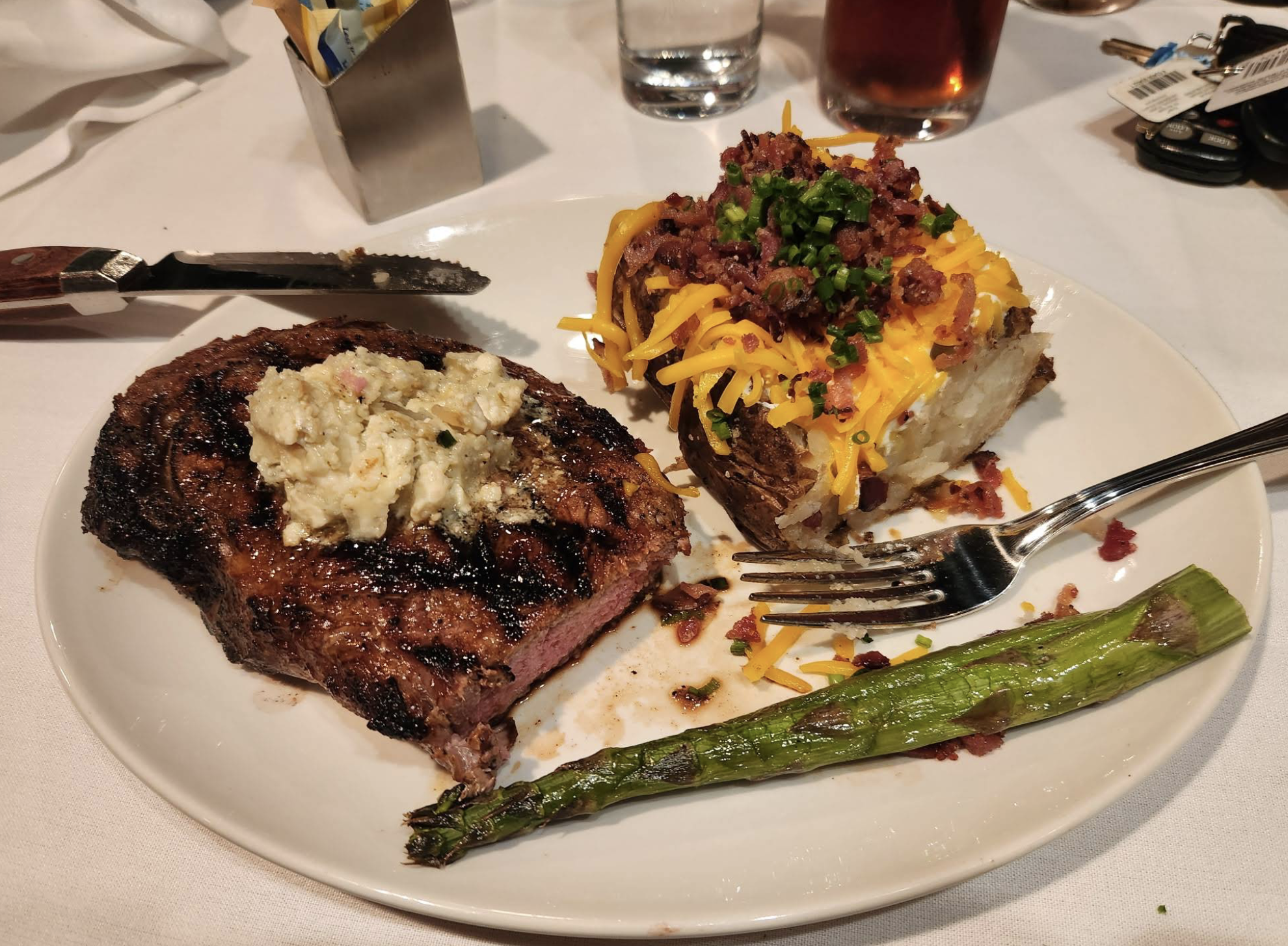 A plate with a grilled steak topped with herb butter, a baked potato loaded with cheese, bacon, and chives, and a grilled asparagus spear, with a fork and knife on a white tablecloth.