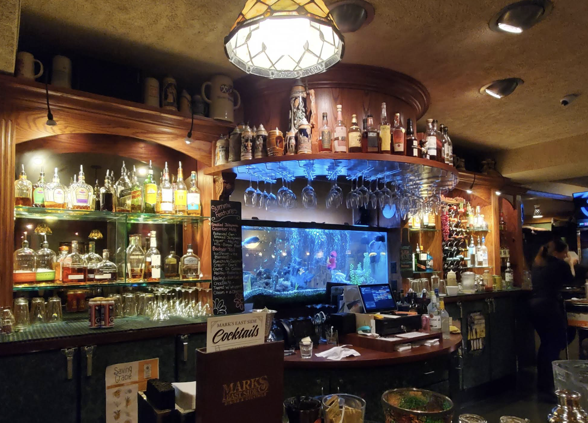 A warmly lit bar with rows of liquor bottles, hanging glasses, and a large aquarium behind the counter; a bartender is blurred in the background. A cocktail menu and bar tools are visible on the counter.