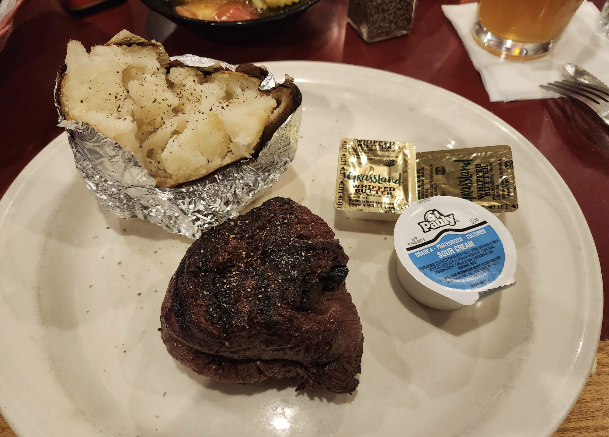 A plate with a baked potato in foil, a grilled beef steak, two butter packets, and a sour cream cup. A drink, napkin, and partially visible bread basket are in the background.