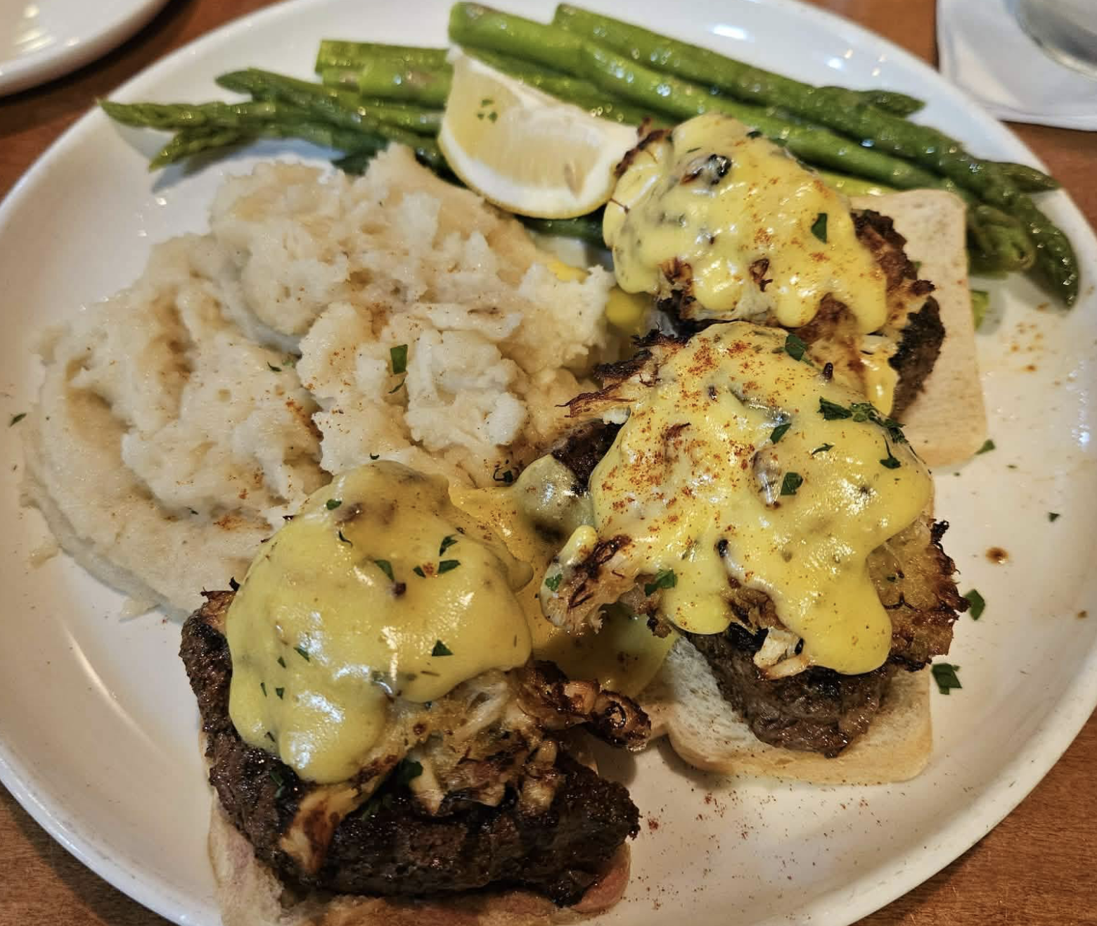 A plate with three pieces of steak topped with a creamy yellow sauce, served on bread slices, alongside mashed potatoes, asparagus, and a lemon wedge.