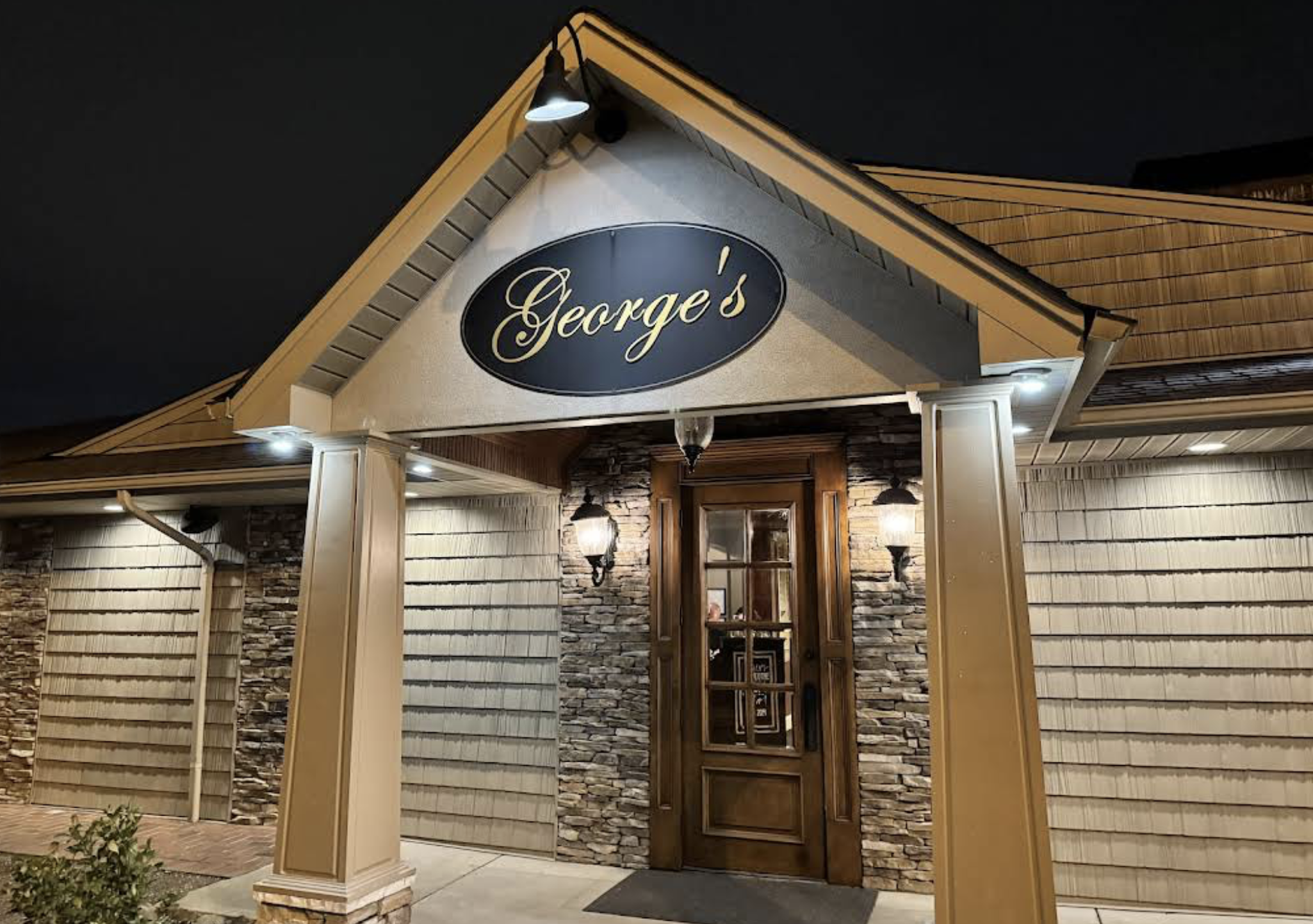 Nighttime view of a restaurant entrance with a sign reading “George’s” above a wooden door, surrounded by stone and shingle siding, lit by several exterior lights.