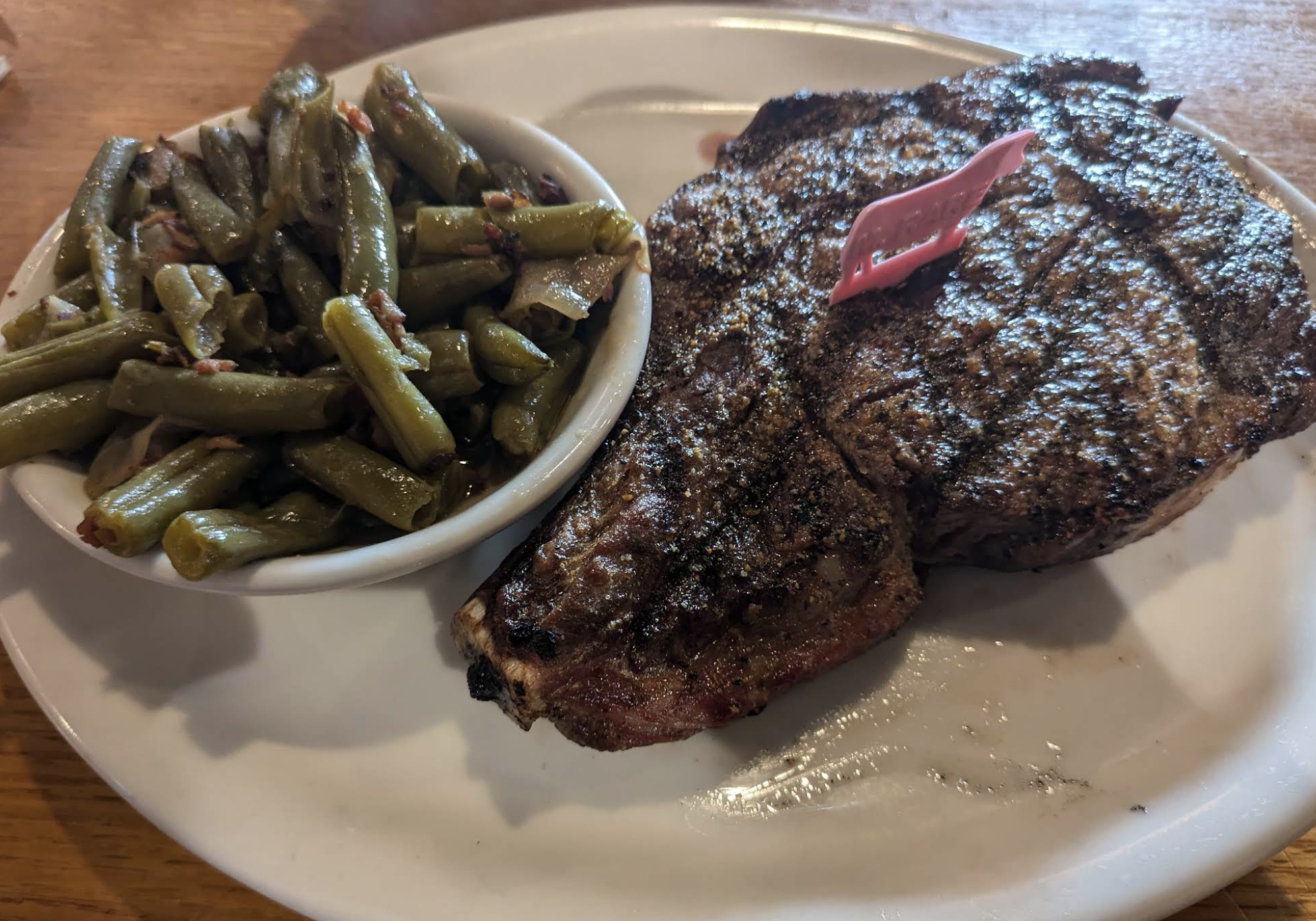 A grilled steak with a pink plastic marker sits on a white plate next to a small bowl of cooked green beans. The plate is on a wooden table.
