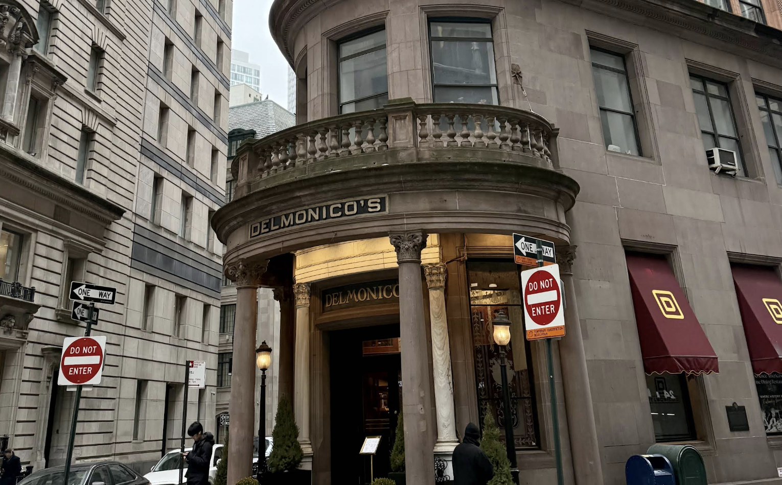A historic stone building with columns and a rounded balcony houses Delmonico’s restaurant. Two "Do Not Enter" street signs are visible near the entrance. People walk on the sidewalk in front.