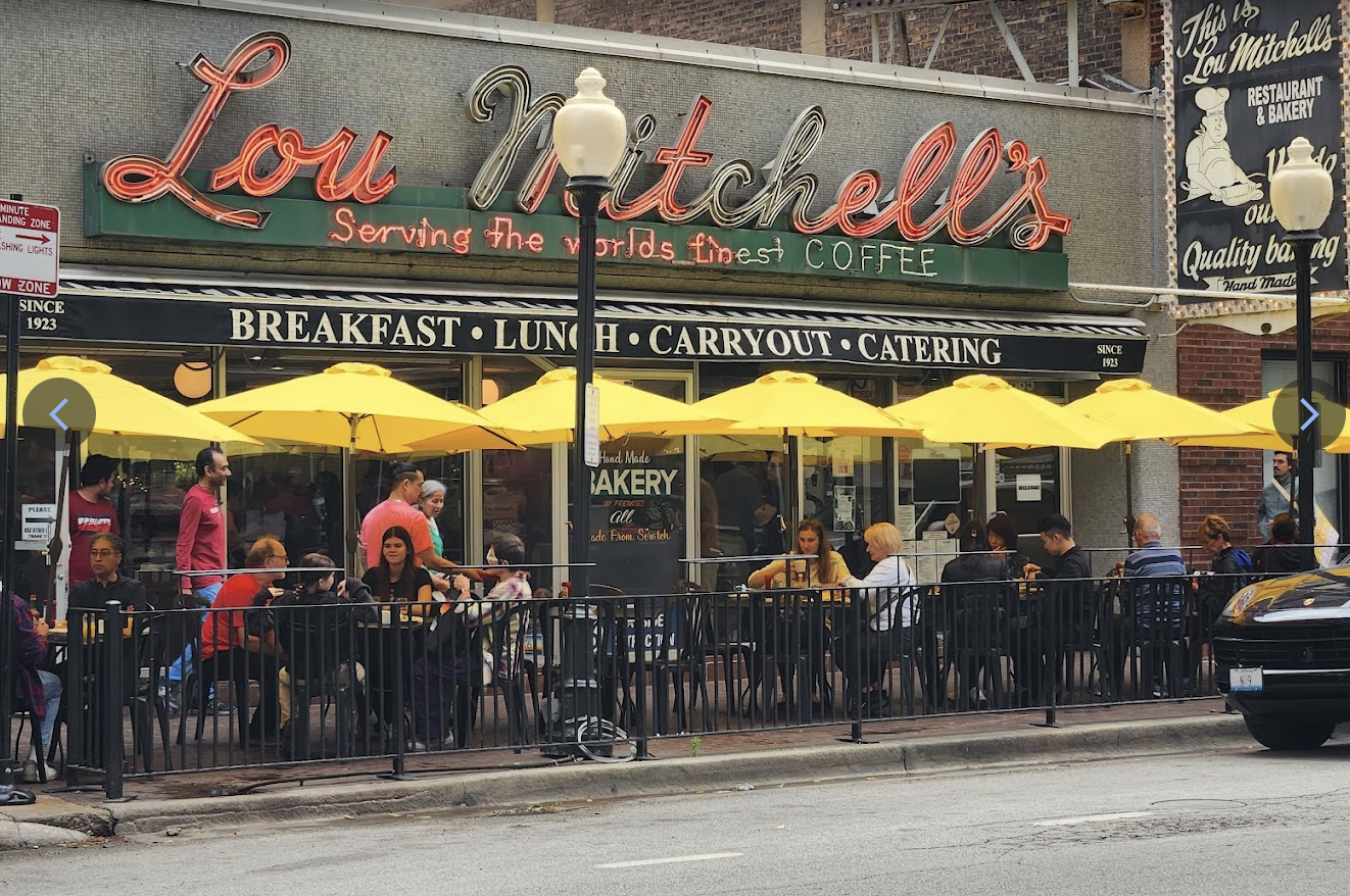 People dine at outdoor tables under yellow umbrellas outside Lou Mitchell’s restaurant, which has a retro sign and advertises breakfast, lunch, carryout, and catering. The street and cars are visible in front.