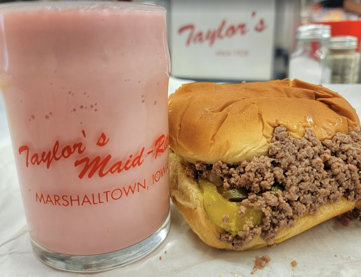 A glass of pink milkshake and a loose-meat sandwich with pickles on a bun are on a table. Both items are from Taylor's Maid-Rite in Marshalltown, Iowa, as shown by the logo on the glass and in the background.