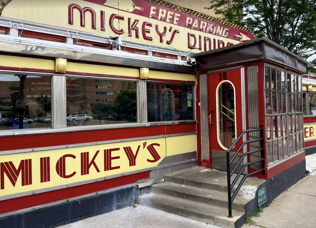 Exterior of Mickey’s Diner, a vintage red and yellow diner with large sign, glass windows, and a small entrance with stairs and railings. Trees and buildings are visible in the background.