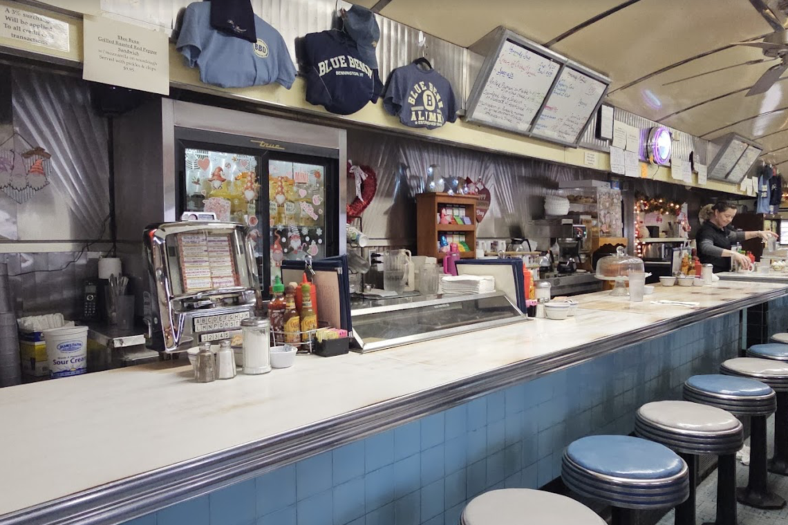 A retro diner interior with a long counter lined with blue and white stools. The counter has condiments, napkin dispensers, and menus. Behind the counter, shelves hold mugs and decor. A server works near the end.