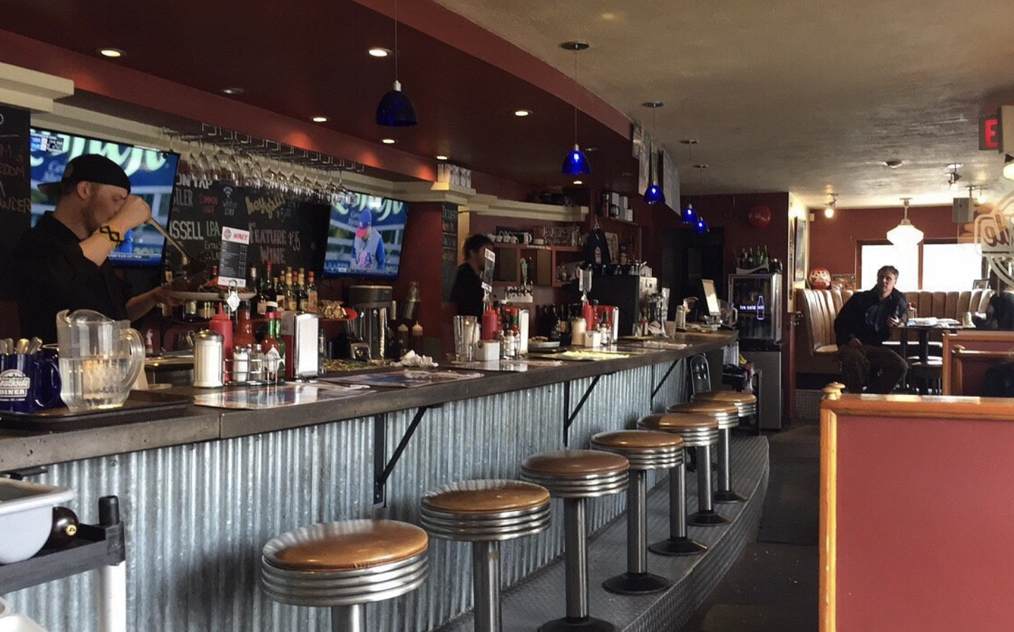 A diner bar with metal stools, a corrugated counter, bottles and condiments, and two staff behind the bar. A few patrons sit at booths in the background under warm lighting. TVs are mounted on the wall.