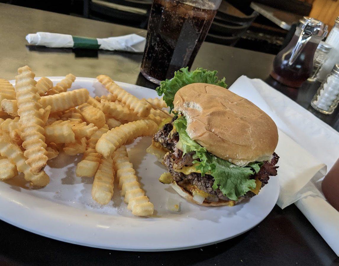 A plate with crinkle-cut French fries and a cheeseburger with lettuce and onions, served next to a glass of soda on a dark table in a restaurant setting.