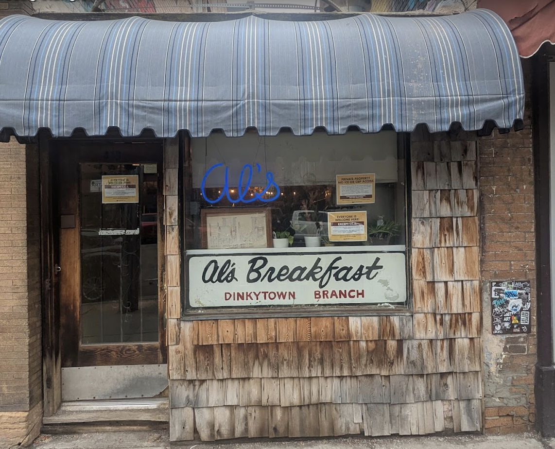 A weathered storefront with a blue-striped awning and wood shingles displays a neon "Al's" sign and a large sign reading "Al's Breakfast Dinkytown Branch" in the window. Notices are posted on the door and inside.