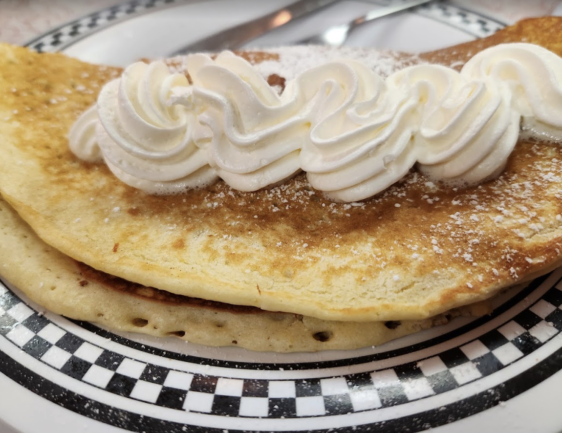 A folded pancake topped with powdered sugar and a swirl of whipped cream, served on a white plate with a black checkered border.