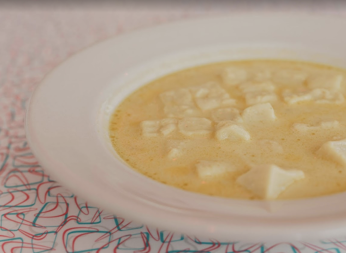 A close-up of a white bowl filled with creamy yellow soup containing small cubes of cheese on a patterned surface with blue and red lines.