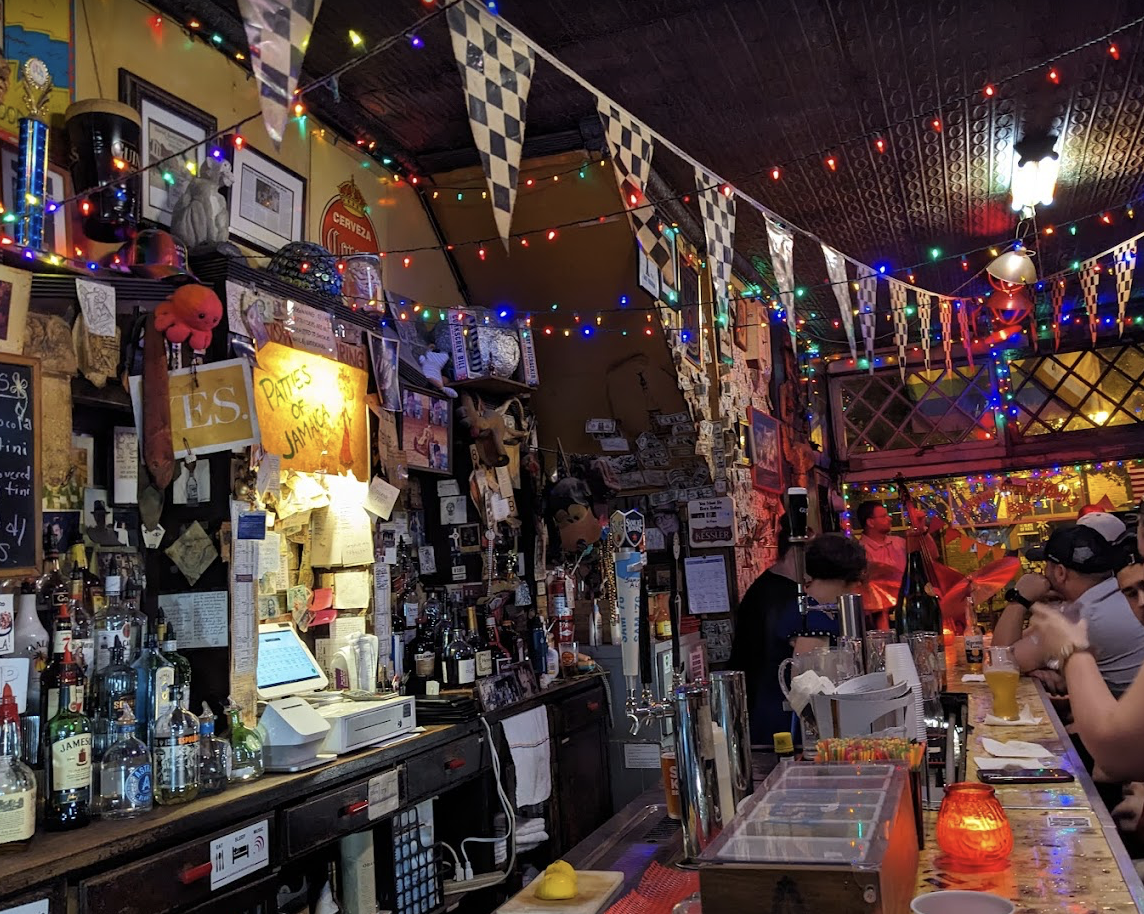 A cozy bar with colorful string lights, checkered flags, and cluttered decor. People sit at the bar, surrounded by bottles, notes, and various eclectic items on the walls and shelves. Warm, festive atmosphere.