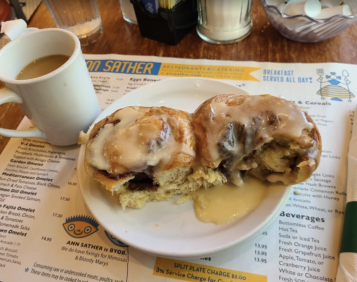 A cinnamon roll covered with white icing is split open on a plate, revealing the inside. A cup of coffee sits to the side, and the meal rests on top of a breakfast menu at a restaurant.