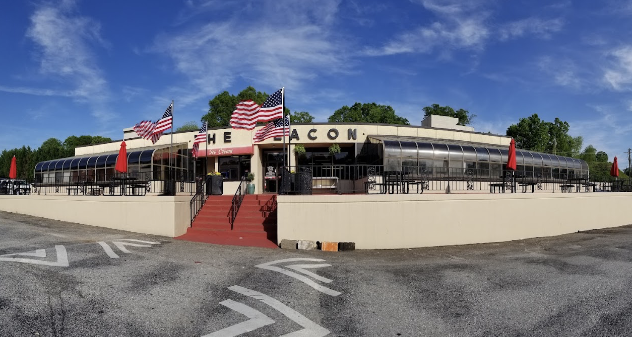A classic diner with a curved glass facade and outdoor seating, displays four American flags in front. Red steps lead to the entrance under a sign that partially reads "THE BEACON." Trees and a blue sky are in the background.