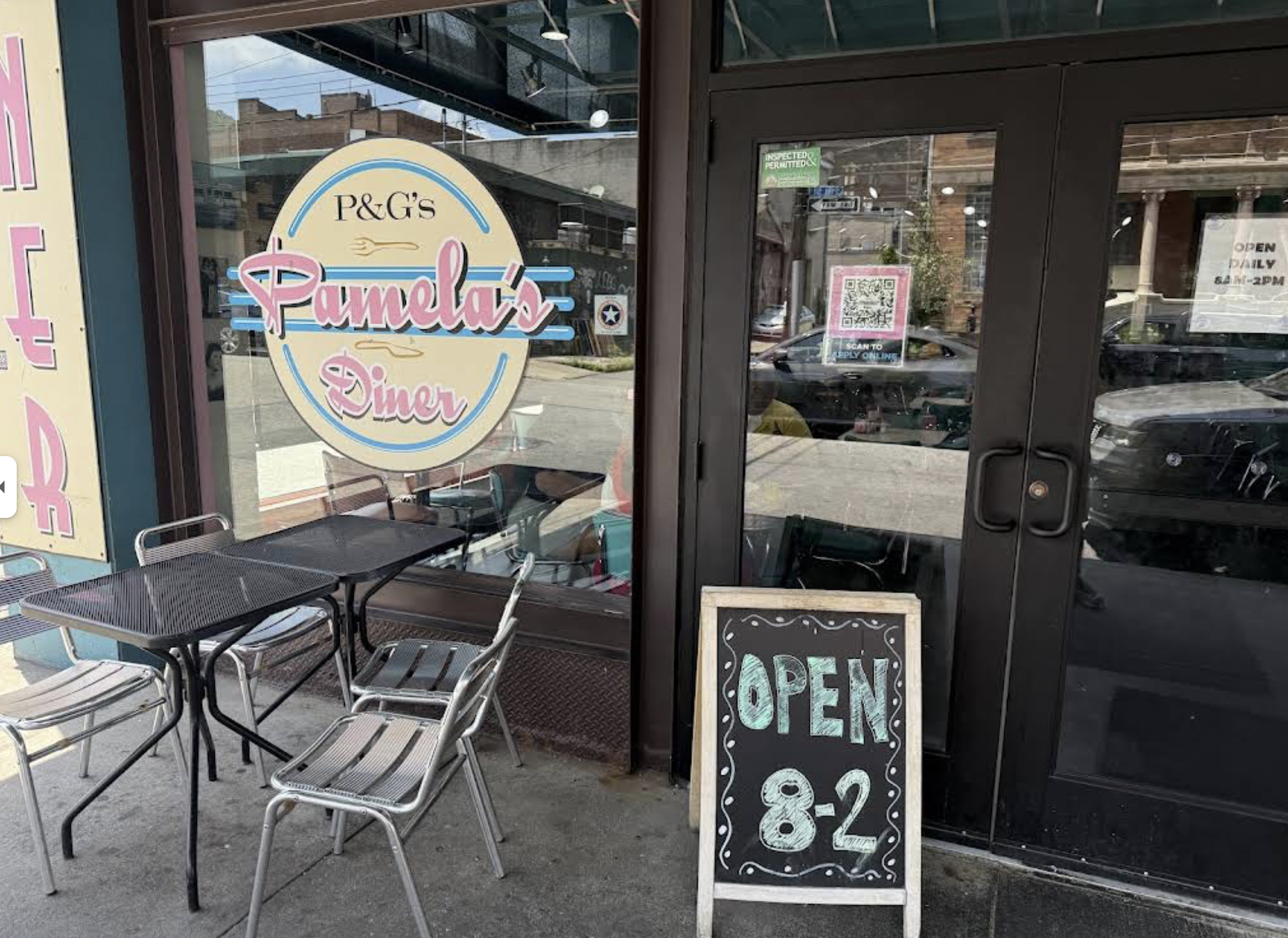 Outdoor seating in front of Pamela’s Diner. A round sign on the window displays the diner's name; a chalkboard sign by the door shows "OPEN 8-2." Empty tables and chairs are set up on the sidewalk.