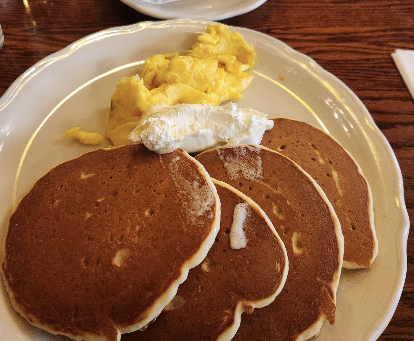 Three golden-brown pancakes topped with a dollop of butter are served alongside a portion of scrambled eggs on a white plate, set on a wooden table.