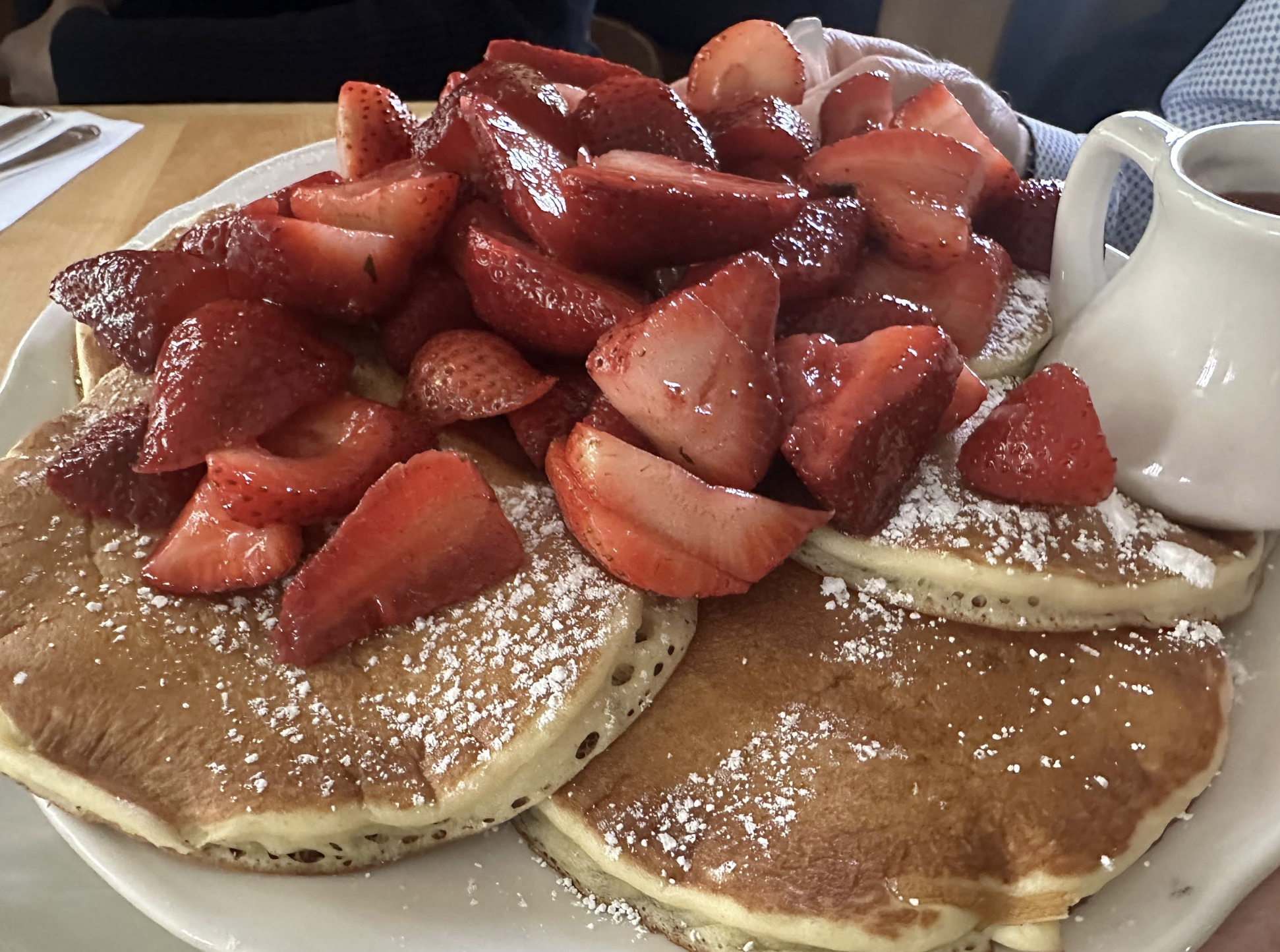 A plate of pancakes topped with sliced strawberries and powdered sugar, with a small white pitcher of syrup on the side.