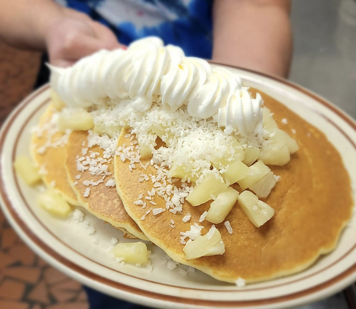 A person holds a plate with three pancakes topped with whipped cream, shredded coconut, and pineapple chunks.