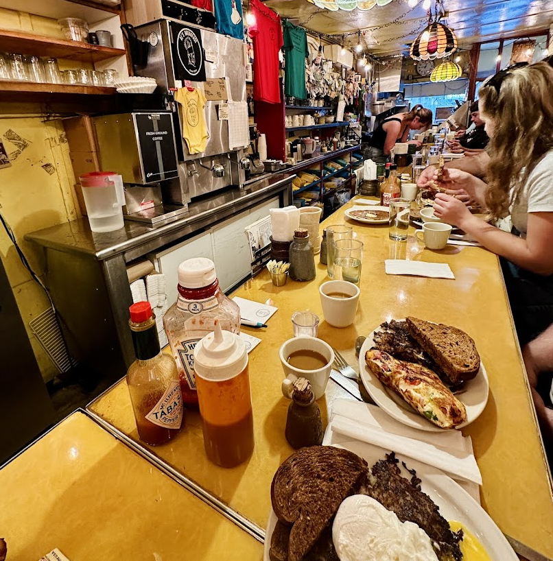 A bustling diner scene with people sitting at a yellow counter, eating breakfast. Plates of toast, eggs, and brisket are in the foreground, and bottles of condiments and coffee cups are on the counter. The atmosphere is cozy and lively.