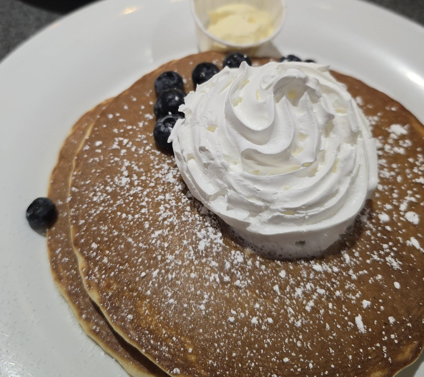 Two pancakes topped with whipped cream, a few blueberries, and a dusting of powdered sugar on a white plate. A small cup of butter is in the background.