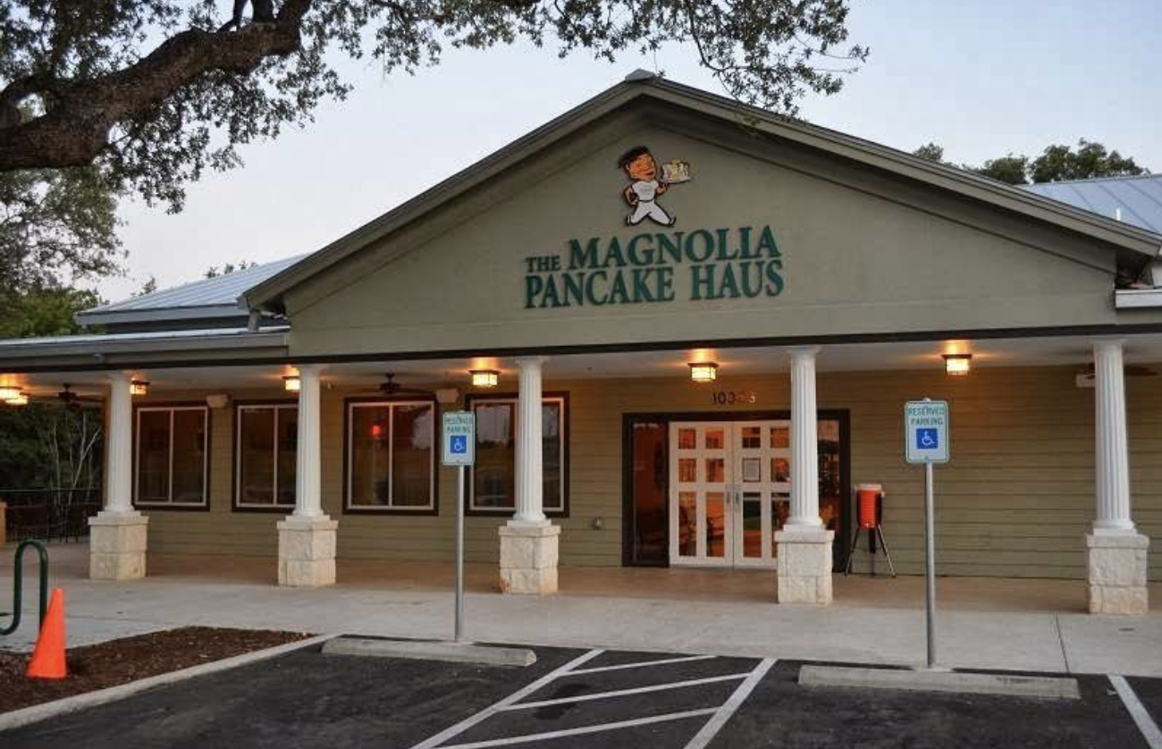 The exterior of The Magnolia Pancake Haus restaurant, featuring a green facade, white pillars, double glass doors, accessible parking spaces, and a sign with a cartoon chef above the entrance.