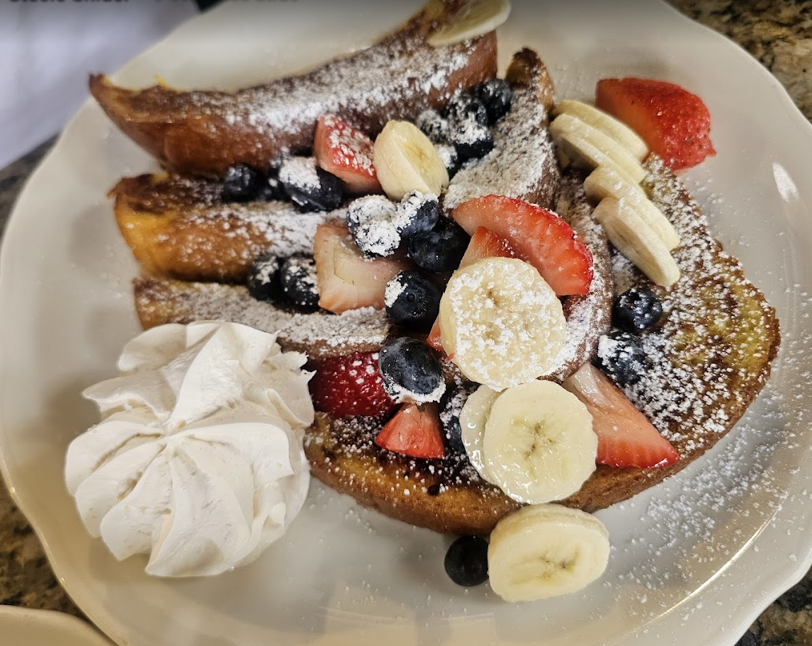 A plate of French toast topped with powdered sugar, sliced bananas, strawberries, blueberries, and a dollop of whipped cream on the side.