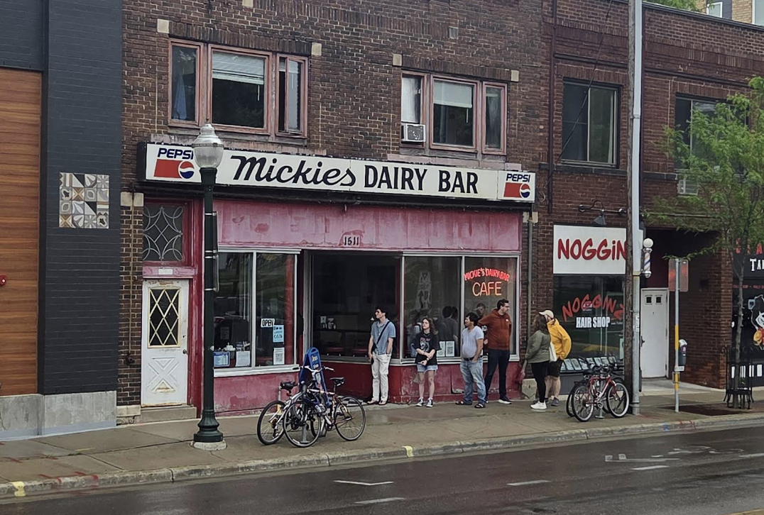 A group of people stand outside Mickies Dairy Bar, a vintage brick building with a faded pink facade and Pepsi signs, on a wet city sidewalk. Bicycles are parked in front, and Noggin hair shop is next door.