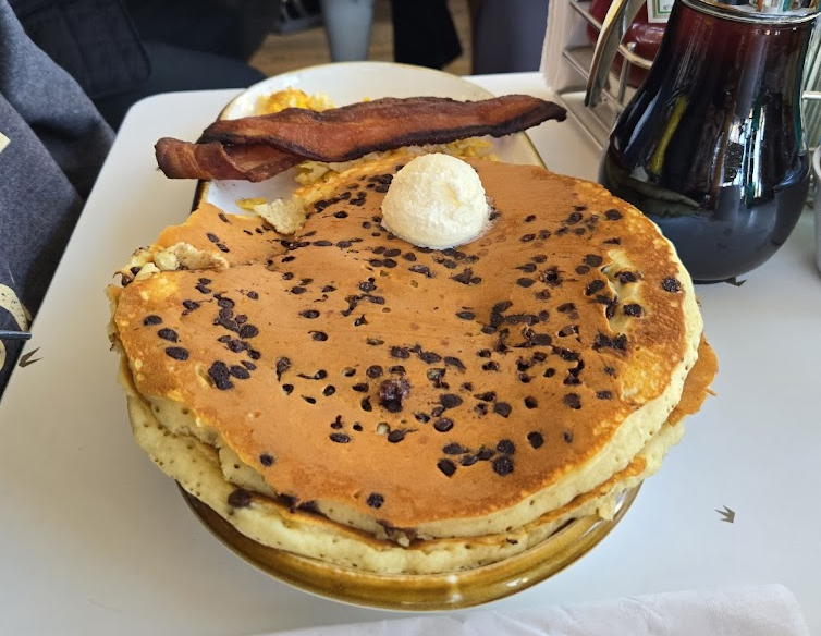 A plate with two chocolate chip pancakes topped with a scoop of butter, a strip of crispy bacon, and scrambled eggs on the side. A bottle of syrup is visible nearby on the table.