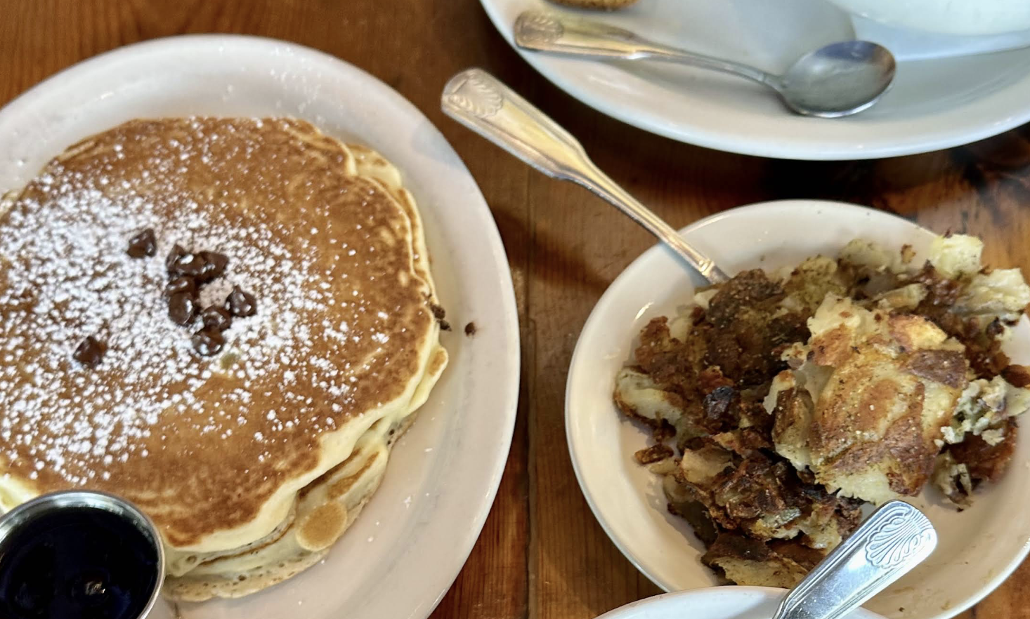 A plate of pancakes topped with powdered sugar and chocolate chips sits beside a bowl of crispy hash browns on a wooden table, with a small cup of syrup and spoons visible nearby.