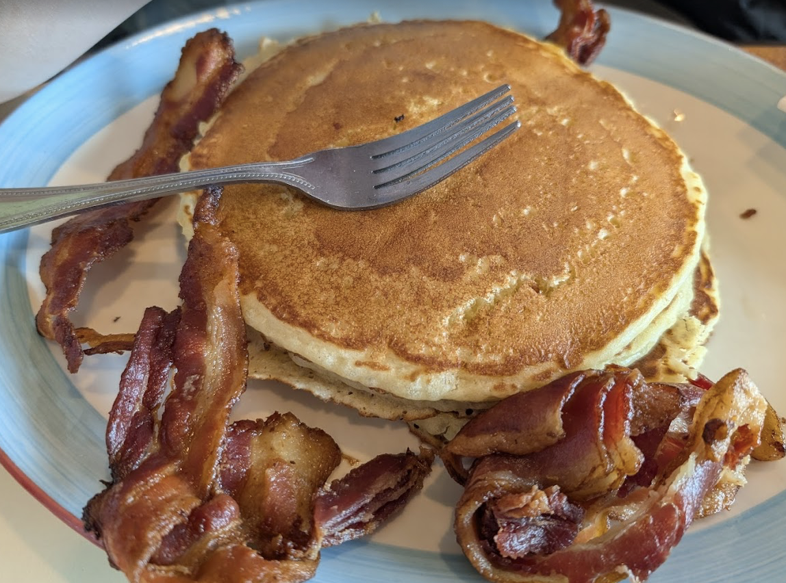 A plate with two golden pancakes stacked, a fork resting on top, and crispy strips of bacon arranged around the pancakes.
