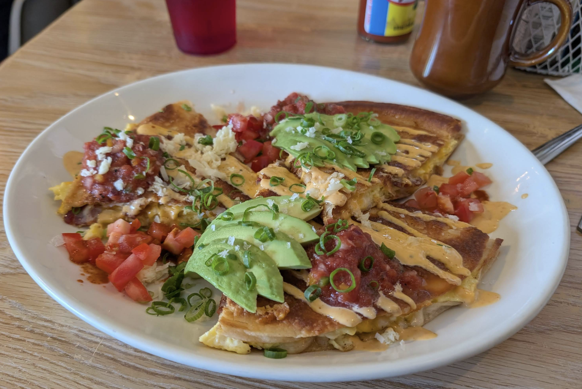 A plate of quesadillas topped with sliced avocado, salsa, diced tomatoes, shredded cheese, and green onions, drizzled with a creamy sauce, on a wooden table with condiments in the background.
