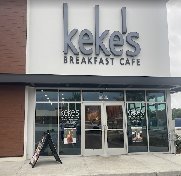 The front entrance of Keke's Breakfast Cafe with large glass doors and windows, a visible street number 8031, and a sidewalk sign displaying menu items outside.