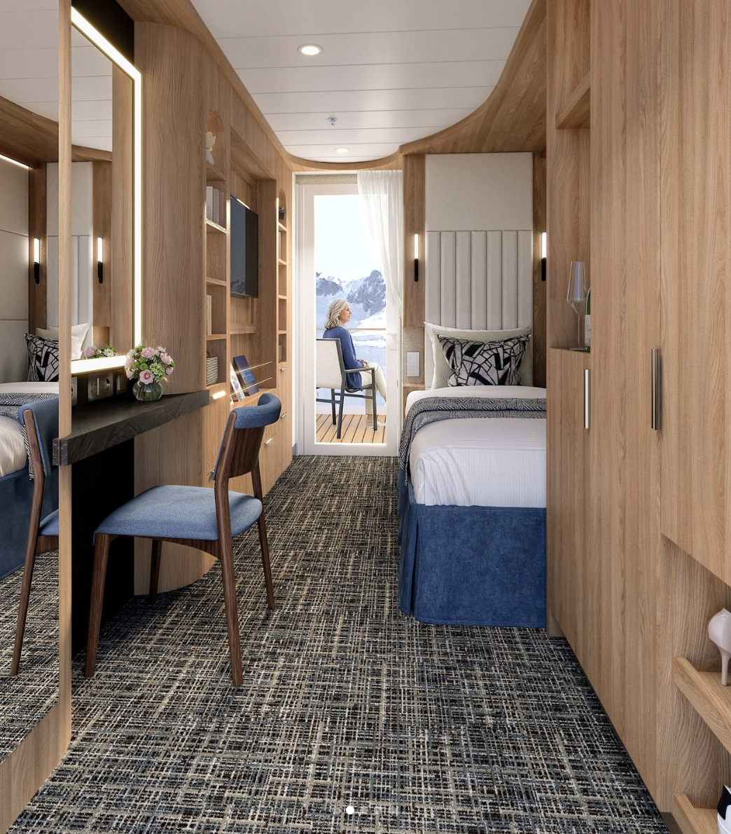 A modern, cozy Best Cruise Line 2026 cabin features a bed, desk, chair, flowers, and large mirror. Through a glass door, a person relaxes on the balcony, soaking in stunning views of snowy mountains and water.