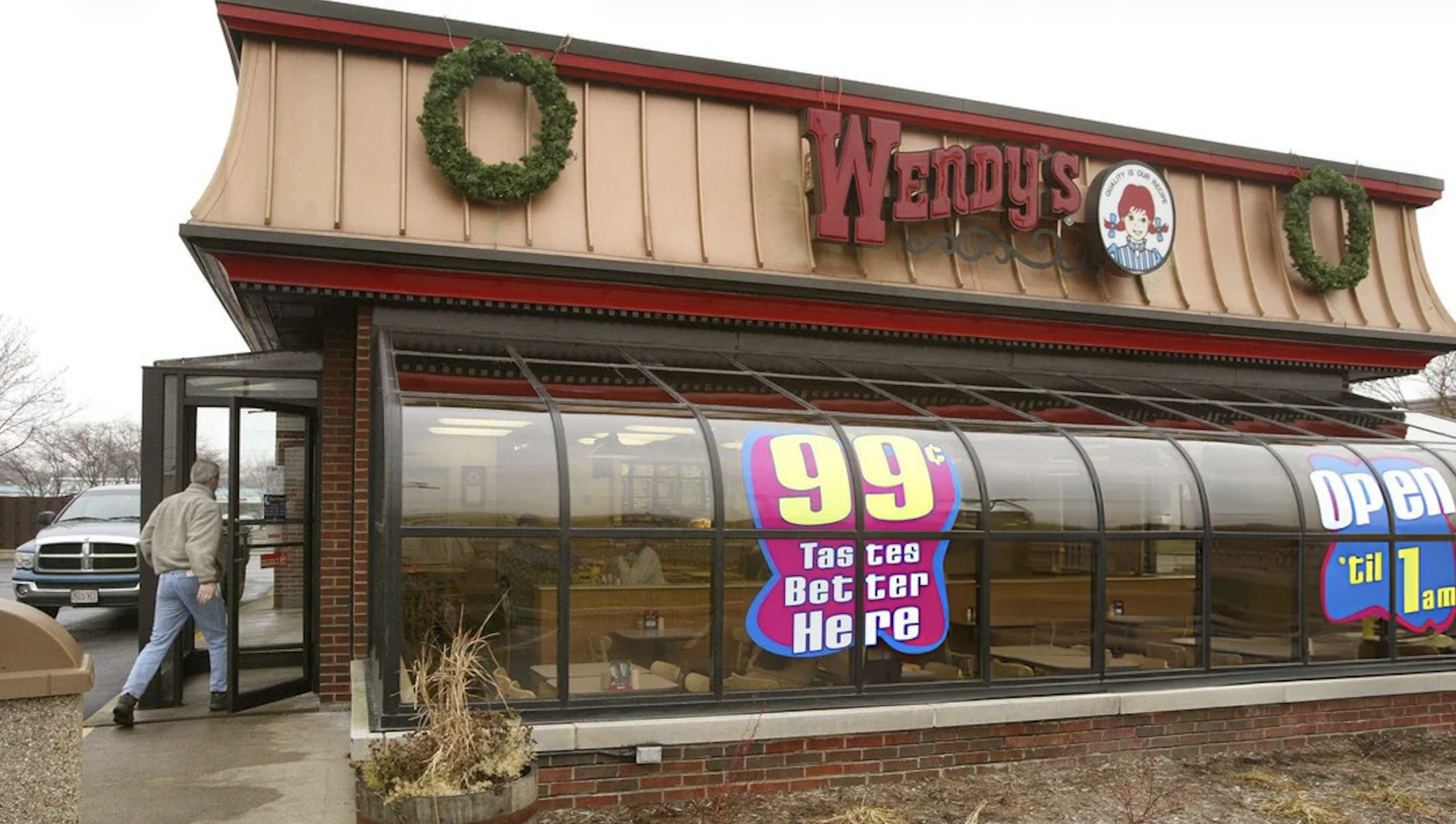 A person enters a Wendy's restaurant with holiday wreaths on the roof. Large window signs promote a 99-cent offer and late hours, with visible empty seating inside—capturing classic charm in Vintage Fast-Food Photos.