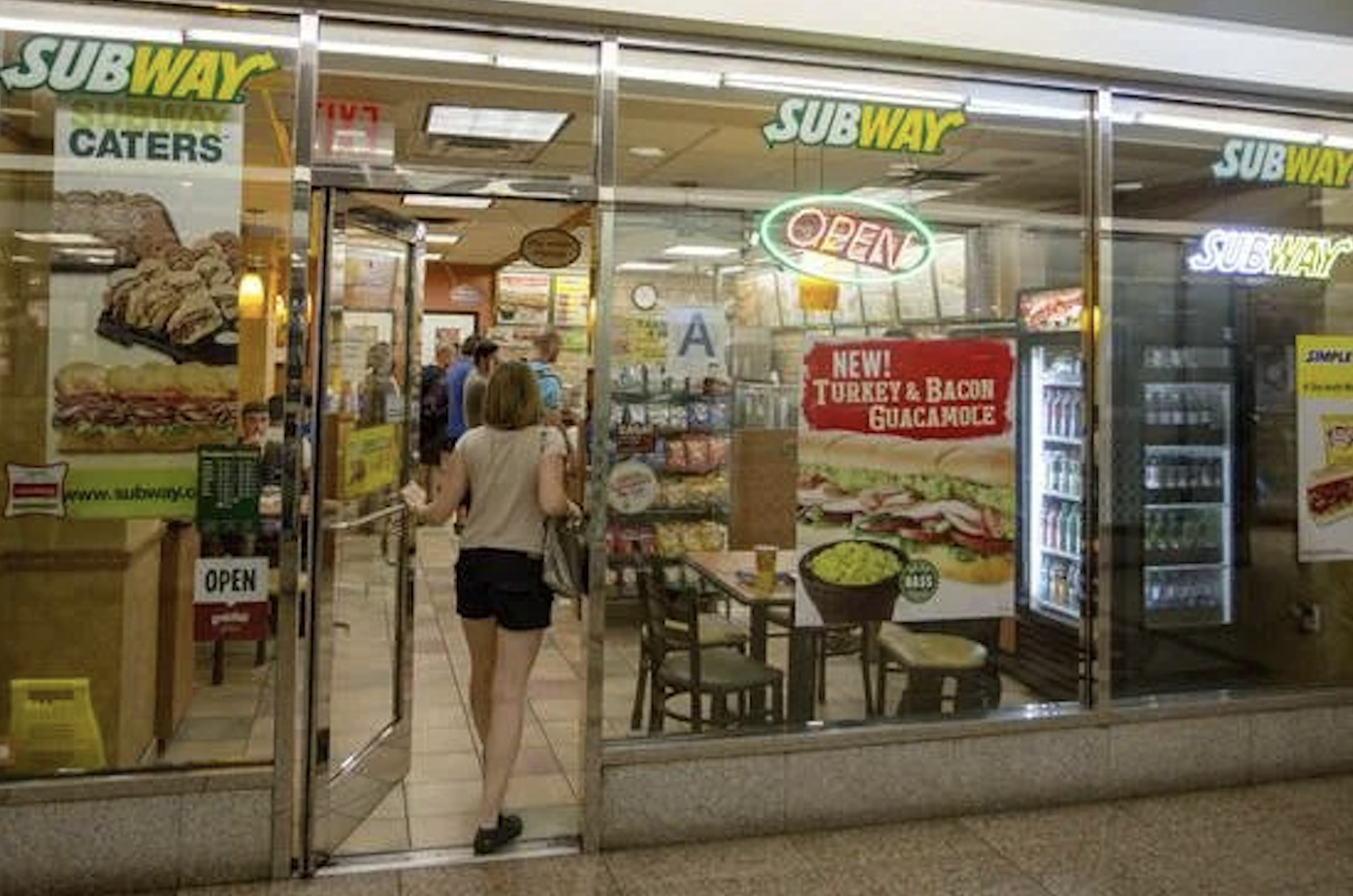 A woman enters a Subway restaurant with glass walls, displaying sandwich posters, an open sign, and a refrigerator with drinks—a scene reminiscent of Vintage Fast-Food Photos. Several customers are inside, and the counter is visible in the background.